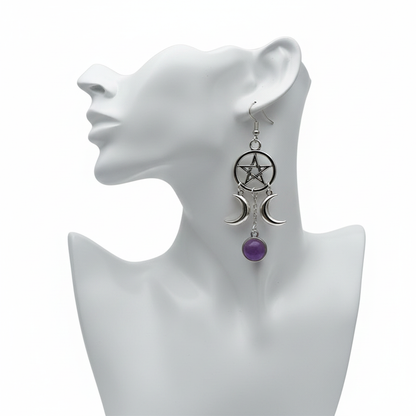 A silver-toned gothic-style dangle earring featuring a pentagram within a circle at the top. Two crescent moons hang from the sides, flanking a central chain that drops down to a round, purple amethyst-style cabochon. The earring is displayed on a white mannequin head against a plain white background.
