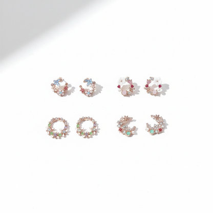 Four pairs of rose gold-tone stud earrings in delicate circular wreath and crescent shapes, featuring pastel blue, pink, and green gemstones with tiny floral accents on a bright white background.