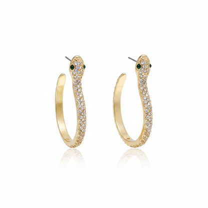 A pair of elegant 18k gold-plated snake hoop earrings featuring shimmering white crystal pavé and striking green emerald-style eyes on a white reflective background.