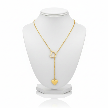 A high-resolution product shot of a gold heart lariat necklace featuring an open-heart slider and a solid heart pendant at the end of a drop chain, displayed on a classic white jewelry bust against a pure white background.