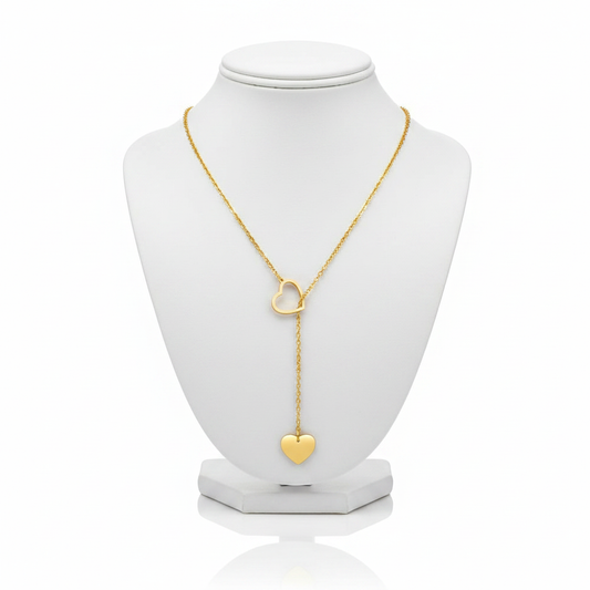 A high-resolution product shot of a gold heart lariat necklace featuring an open-heart slider and a solid heart pendant at the end of a drop chain, displayed on a classic white jewelry bust against a pure white background.