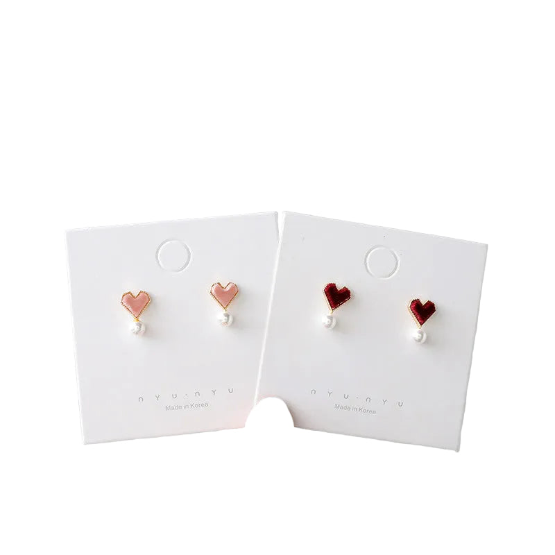 Two pairs of heart-shaped stud earrings displayed on white "nyunyu" branded cards. One pair features light pink hearts with a gold border and a small white pearl dangling below, while the other pair features deep burgundy velvet hearts with similar pearl accents. Both are labeled "Made in Korea."