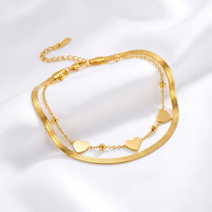 MR BATZ 18k gold-plated layered anklet featuring a sleek herringbone chain and a delicate bead-and-heart charm link, presented in a warm luxury art gallery aesthetic.