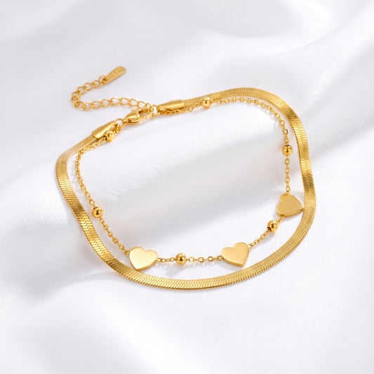 MR BATZ 18k gold-plated layered anklet featuring a sleek herringbone chain and a delicate bead-and-heart charm link, presented in a warm luxury art gallery aesthetic.