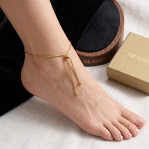 A sleek MR BATZ gold snake chain anklet with an adjustable lariat drop, styled on a foot with a matching gold toe ring in an editorial art gallery aesthetic.