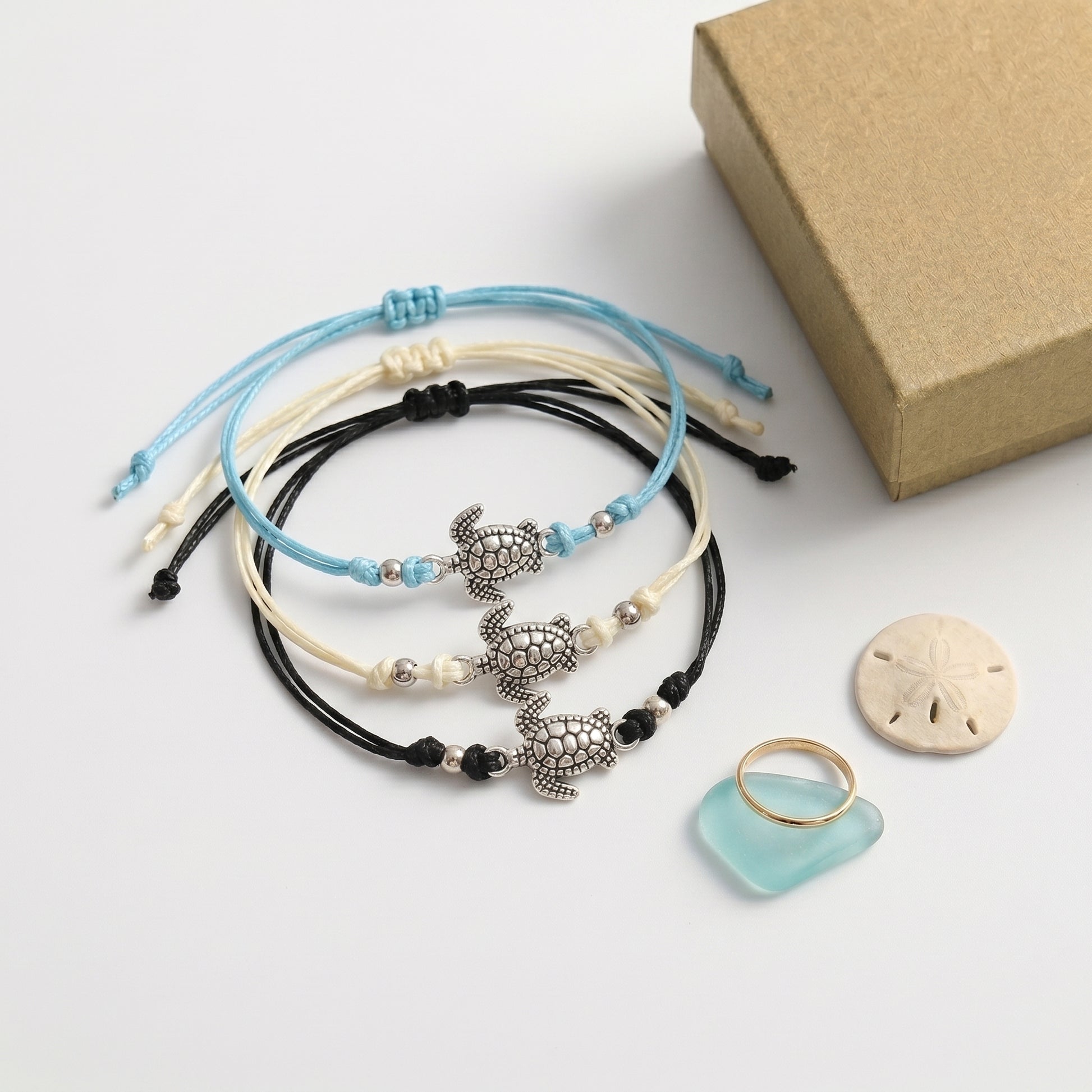 MR BATZ handcrafted adjustable cord bracelets featuring silver turtle charms in blue, cream, and black, displayed with a gold ring and sea glass in a warm luxury art gallery style.