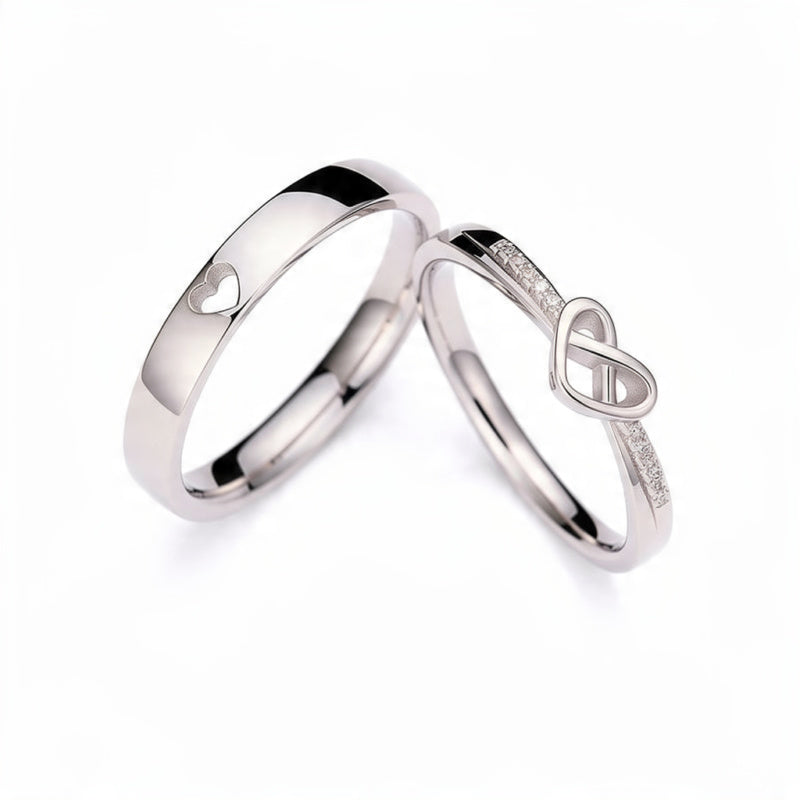 Two adjustable platinum tone couple rings on white background, one band with heart cutout design and one with infinity symbol and crystal stones, matching his and hers promise rings set