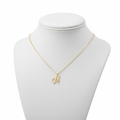 A gold-toned necklace featuring a delicate pendant that blends a script-style initial "A" with an integrated open-heart design. The pendant is suspended from a fine gold cable chain and displayed on a white jewelry mannequin bust against a pure white background.