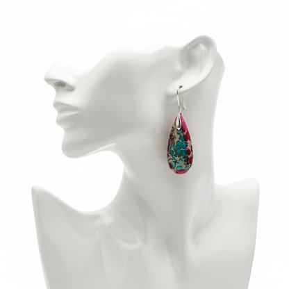 A pair of statement dangle earrings featuring large teardrop-shaped stones with a vibrant, marbled pattern of pink, blue, cream, and tan. Each stone is set in a thin gold-toned metal frame and hangs from a simple gold French hook. The earrings are displayed on a white mannequin head against a clean, neutral background.