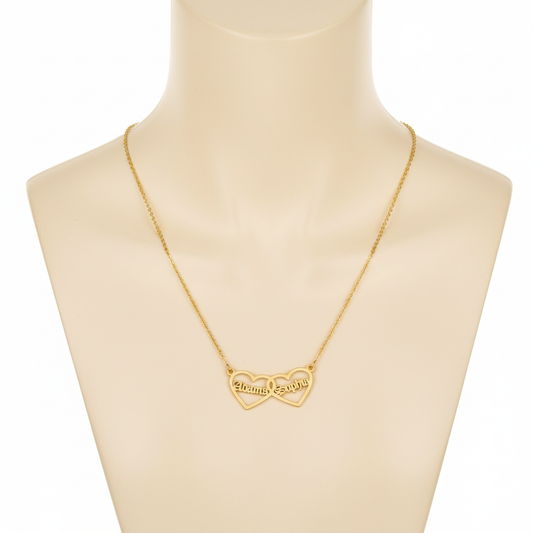 A gold-toned necklace featuring two interlocking open-heart pendants, displayed on a beige mannequin bust. Each heart is personalized with a name in a Gothic-style font: "Alonma" on the left and "Sophia" on the right. The pendant is attached to a fine gold cable chain.