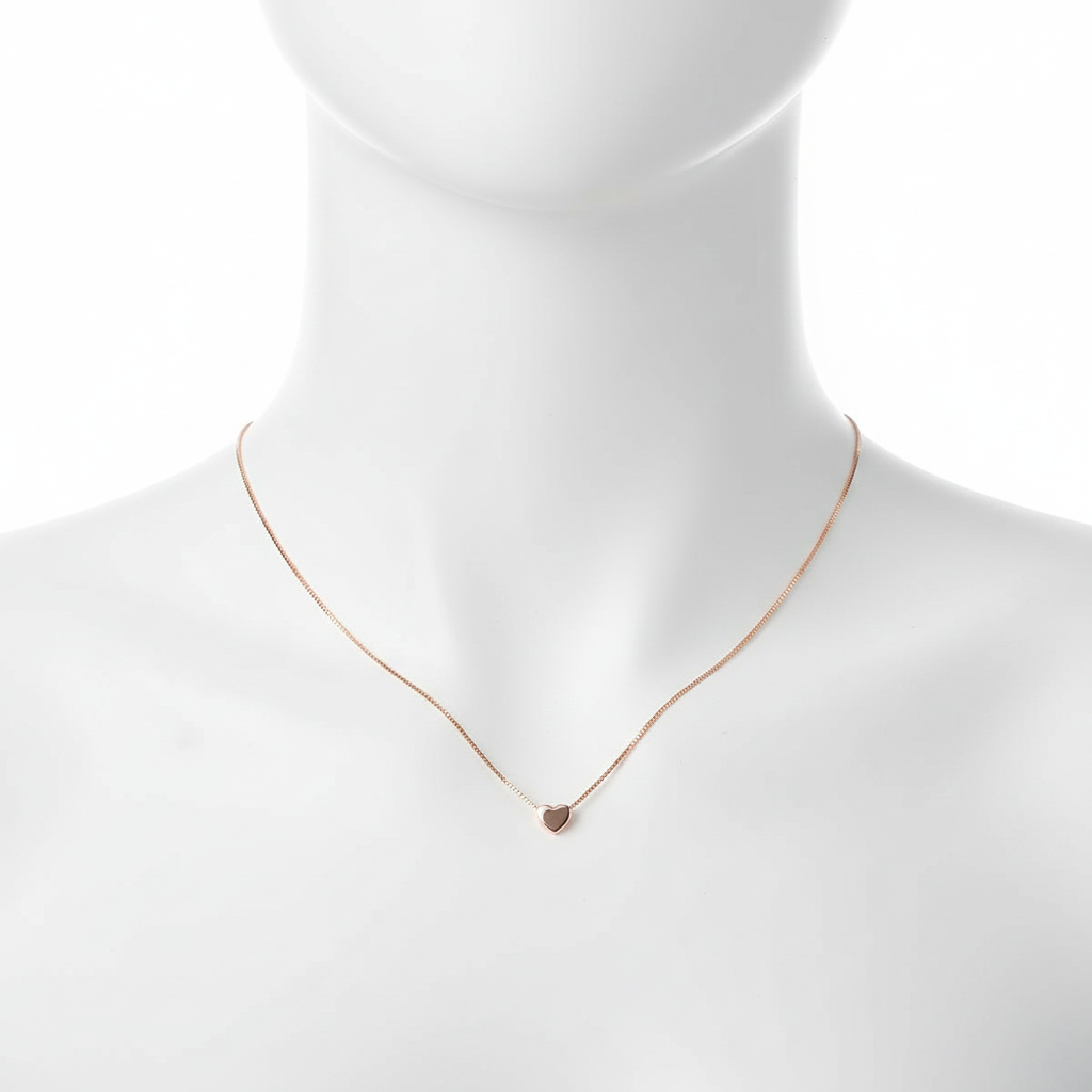 A close-up, high-resolution shot of a dainty rose gold necklace featuring a tiny, polished heart pendant on a thin box chain, displayed on a clean white jewelry mannequin against a pure white background.
