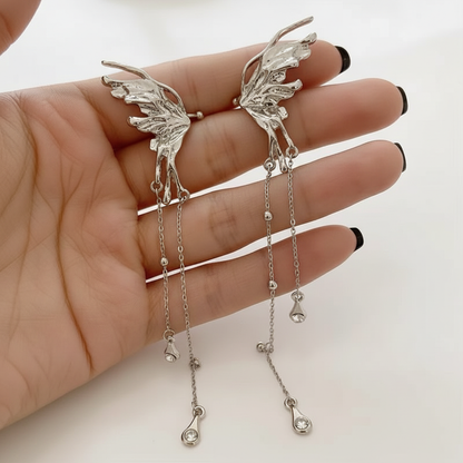 A pair of intricate silver butterfly wing ear cuffs with long dangling chain tassels and crystal teardrop charms held in a hand.