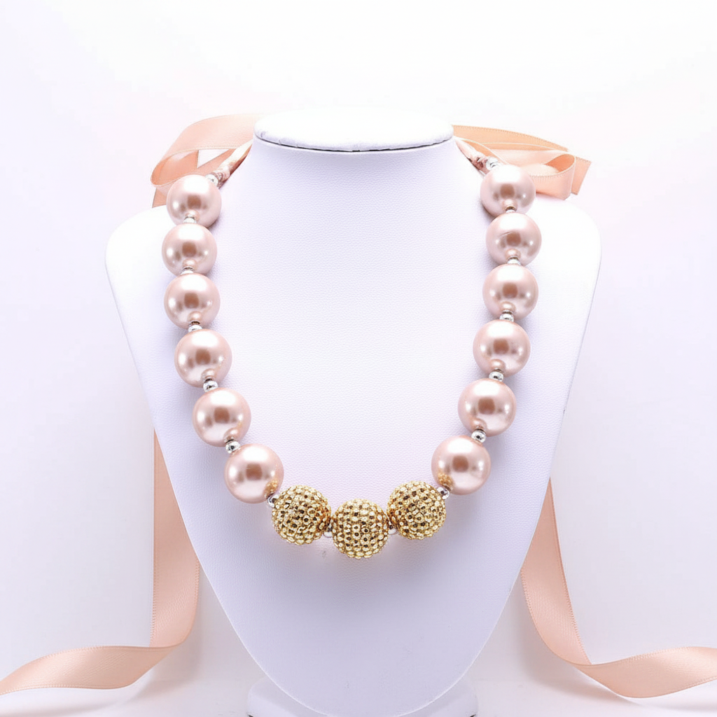 A bold statement necklace featuring oversized blush-toned pearls and textured gold-finish focal beads, finished with a soft satin ribbon tie for a sophisticated, high-impact editorial look.
