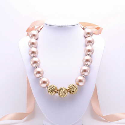 A bold statement necklace featuring oversized blush-toned pearls and textured gold-finish focal beads, finished with a soft satin ribbon tie for a sophisticated, high-impact editorial look.