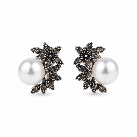 Pair of antique silver-tone stud earrings featuring an intricate floral leaf design set with dark grey marcassite stones and a large, high-luster white pearl, displayed on a clean white background.