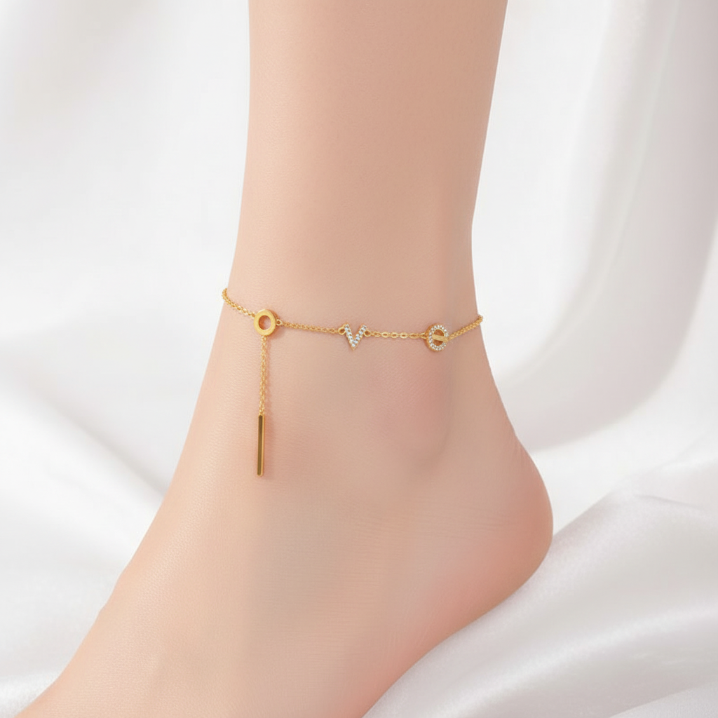Dainty gold chain anklet featuring crystalline letter charms spelling LOVE with a hanging bar pendant on a model's ankle
