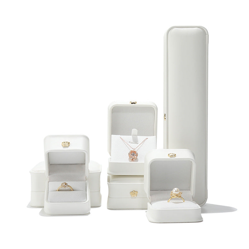 A sophisticated set of white leather jewelry boxes with gold crown emblems, showcasing a diamond ring, a pearl ring, and a rose gold necklace on a clean, high-contrast background.