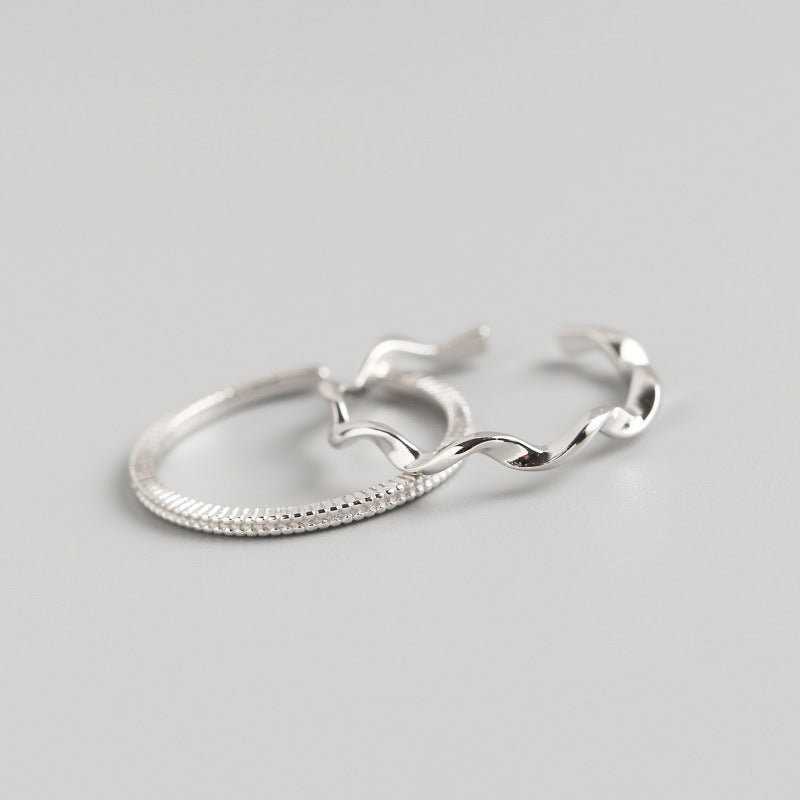 High-angle product photography of two silver-tone rings on a charcoal grey surface; one ring features a consistent beaded texture and the other a polished, undulating wave design with an open back.